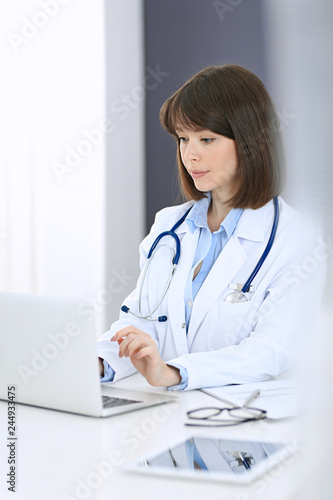 Doctor typing on laptop computer while sitting at the white table in hospital office. Physician at work. Medicine and healthcare concept