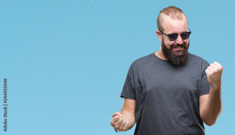 Naklejka premium Young caucasian hipster man wearing sunglasses over isolated background celebrating surprised and amazed for success with arms raised and open eyes. Winner concept.