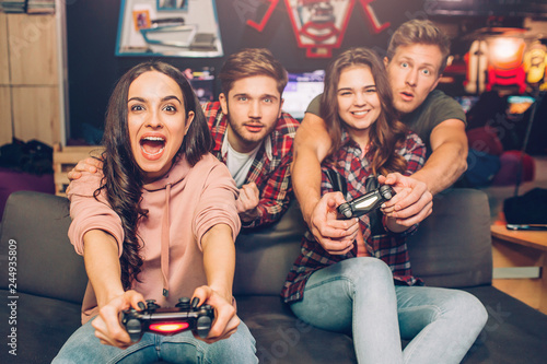 Two young women play intence gam in playing room. THey hold gamepads and look straight forward. Guys try to help them. photo