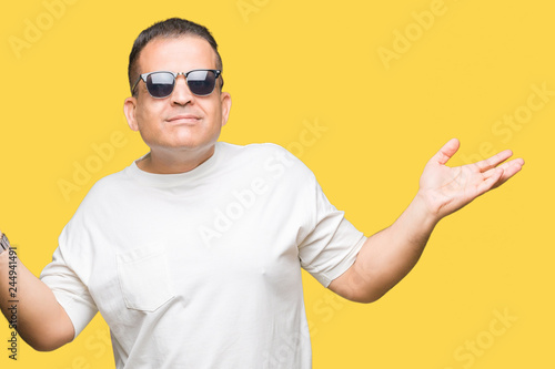 Middle age arab man wearig white t-shirt and sunglasses over isolated background clueless and confused expression with arms and hands raised. Doubt concept.