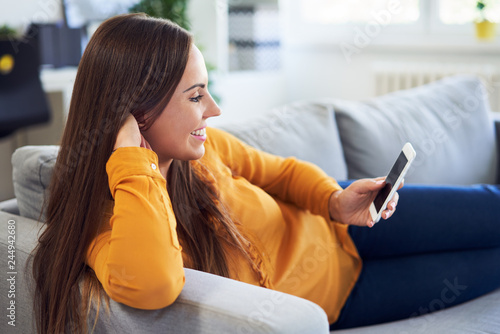 Cheerful woman lying on couch at home and looking at smartphone