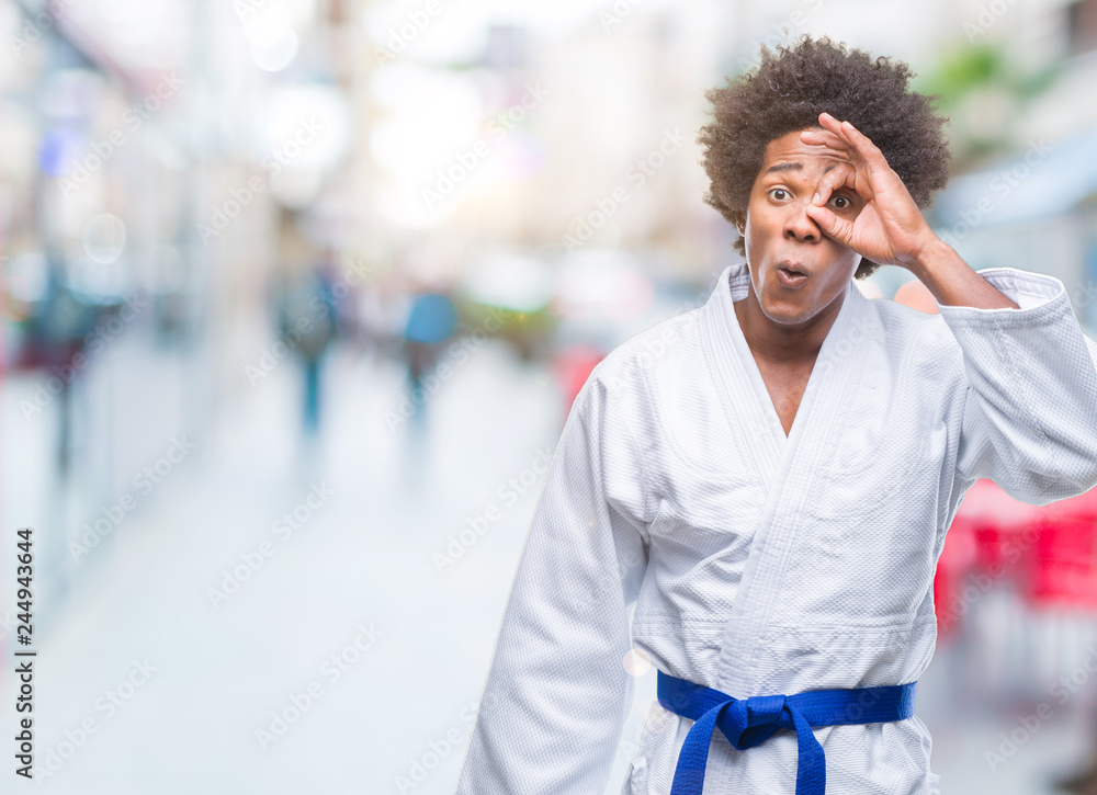 Obraz premium Afro american man wearing karate kimono over isolated background doing ok gesture shocked with surprised face, eye looking through fingers. Unbelieving expression.