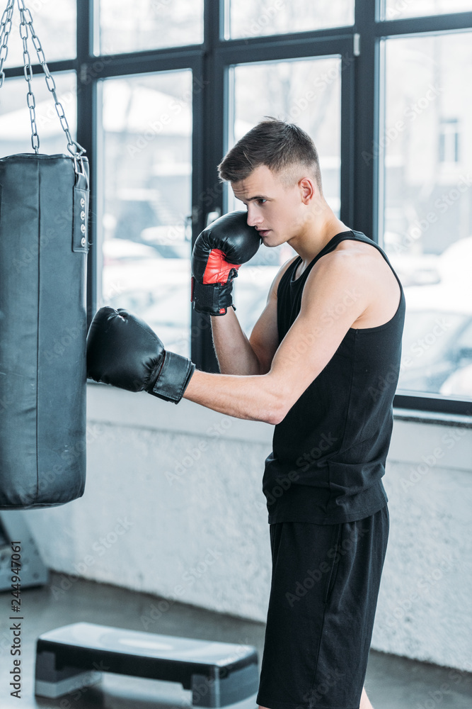 side view of concentrated young man in boxing gloves exercising with punching bag in gym
