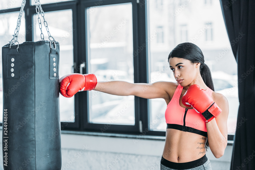 beautiful african american woman in boxing gloves training with punching bag in gym