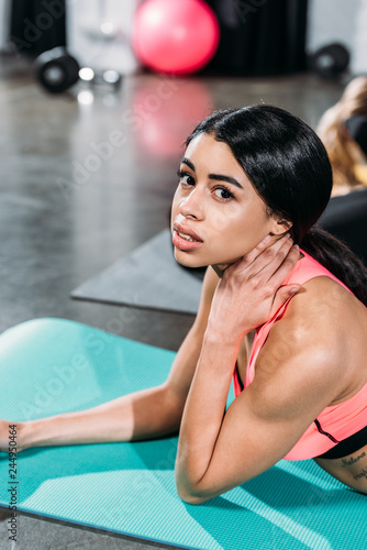 young african american woman touching neck and looking at camera while lying on yoga mat in gym