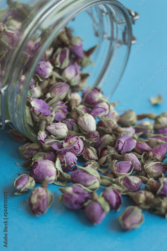 Fototapeta premium Dried rose flowers in a glass jar. Herbal tea on blue background, vertical photo