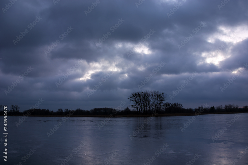 frozen lake, trees on the shore and rainy clouds in the sky