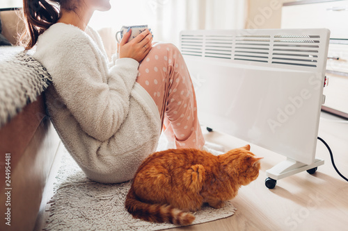 Using heater at home in winter. Woman warming and drinking tea with cat. Heating season.