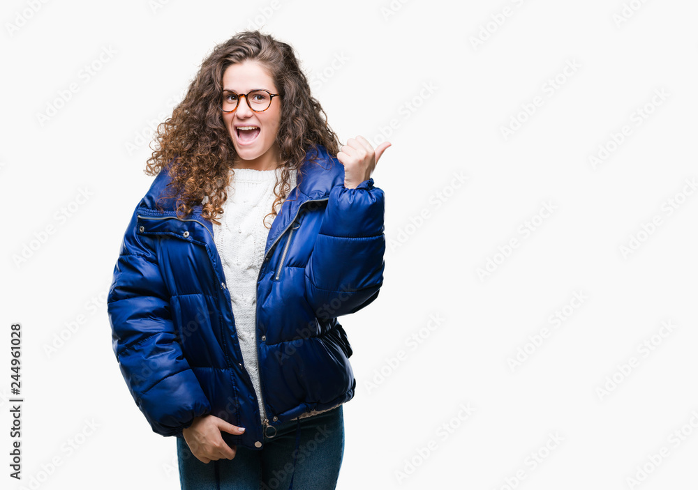 Beautiful young brunette curly hair girl wearing winter coat, glasses and sweater over isolated background smiling with happy face looking and pointing to the side with thumb up. Beautiful young brunette curly hair girl wearing winter coat, glasses and sweater over isolated background smiling with happy face looking and pointing to the side with thumb up.