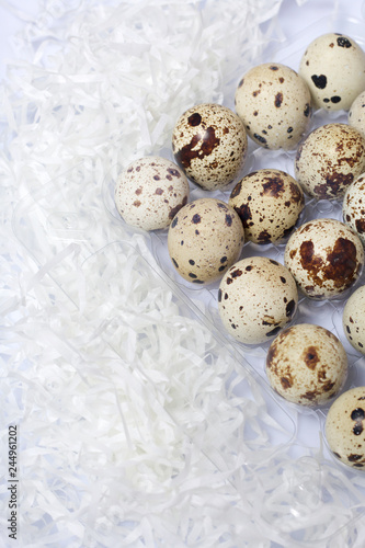 Quail eggs. They lie in a plastic container. Near paper chips. On a white background.