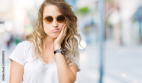 Beautiful young blonde woman wearing sunglasses over isolated background thinking looking tired and bored with depression problems with crossed arms.