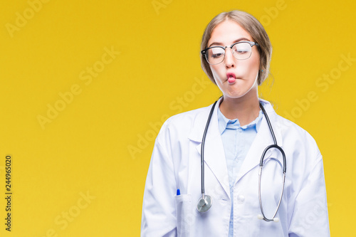 Beautiful young blonde doctor woman wearing medical uniform over isolated background making fish face with lips  crazy and comical gesture. Funny expression.