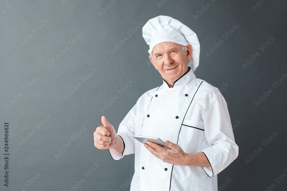 Senior chef studio standing isolated on gray holding digital tablet thumb up satisfied with application Senior chef studio standing isolated on gray holding digital tablet thumb up satisfied with application