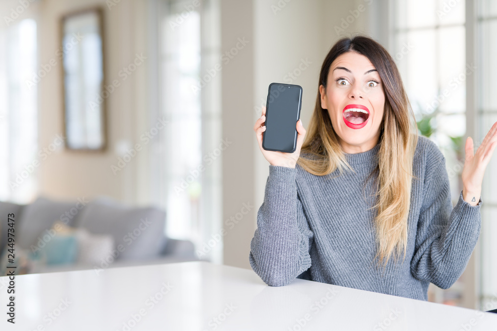 Young beautiful woman showing smartphone screen at home very happy and excited, winner expression celebrating victory screaming with big smile and raised hands
