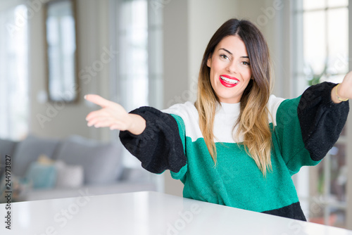Young beautiful woman wearing winter sweater at home looking at the camera smiling with open arms for hug. Cheerful expression embracing happiness. © Krakenimages.com