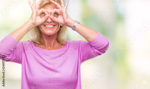 Middle age blonde woman over isolated background doing ok gesture like binoculars sticking tongue out, eyes looking through fingers. Crazy expression.