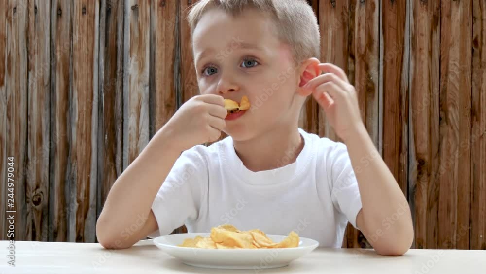 A child eats potato chips with pleasure against the background of a wooden wall. The boy is pleased with the food and shows a thumb up. A child eats potato chips with pleasure against the background of a wooden wall. The boy is pleased with the food and shows a thumb up.