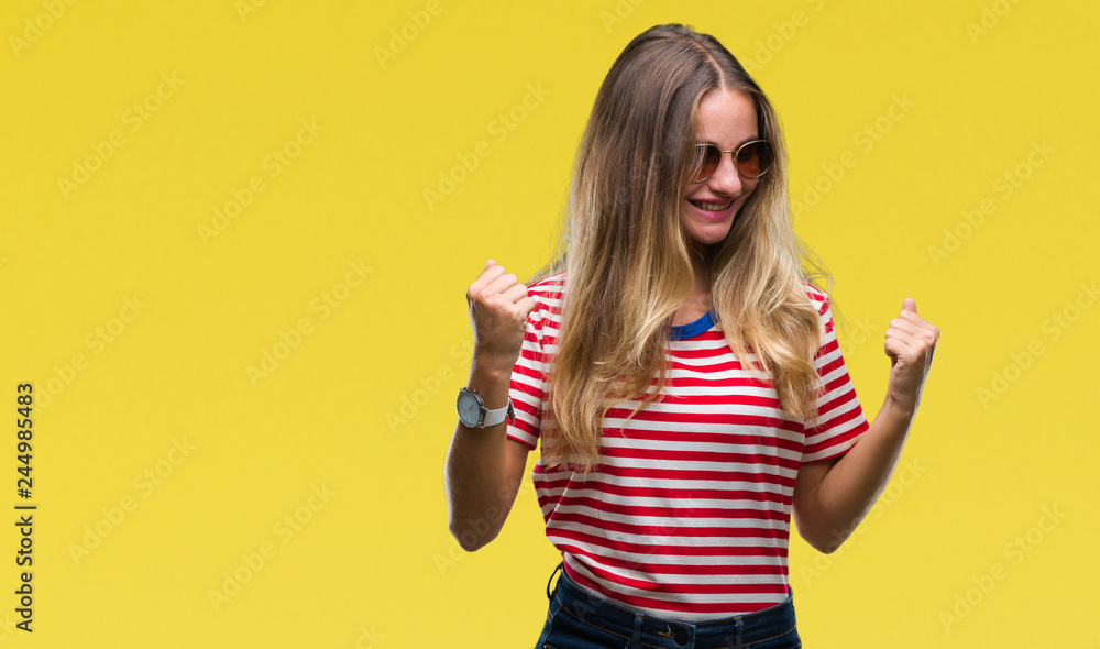 Young beautiful blonde woman wearing sunglasses over isolated background very happy and excited doing winner gesture with arms raised, smiling and screaming for success. Celebration concept. Young beautiful blonde woman wearing sunglasses over isolated background very happy and excited doing winner gesture with arms raised, smiling and screaming for success. Celebration concept.