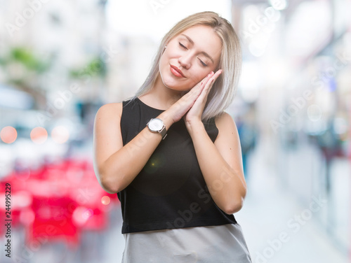 Young blonde woman over isolated background sleeping tired dreaming and posing with hands together while smiling with closed eyes.