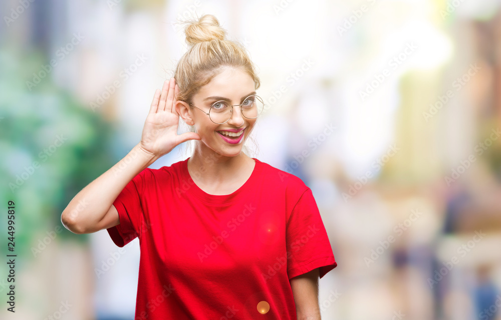 Young beautiful blonde woman wearing red t-shirt and glasses over isolated background smiling with hand over ear listening an hearing to rumor or gossip. Deafness concept. Young beautiful blonde woman wearing red t-shirt and glasses over isolated background smiling with hand over ear listening an hearing to rumor or gossip. Deafness concept.
