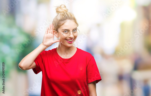 Fototapeta Naklejka Na Ścianę i Meble -  Young beautiful blonde woman wearing red t-shirt and glasses over isolated background smiling with hand over ear listening an hearing to rumor or gossip. Deafness concept.