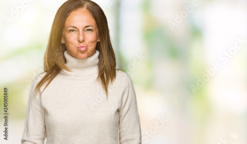 Beautiful middle age adult woman wearing winter sweater over isolated background puffing cheeks with funny face. Mouth inflated with air, crazy expression.