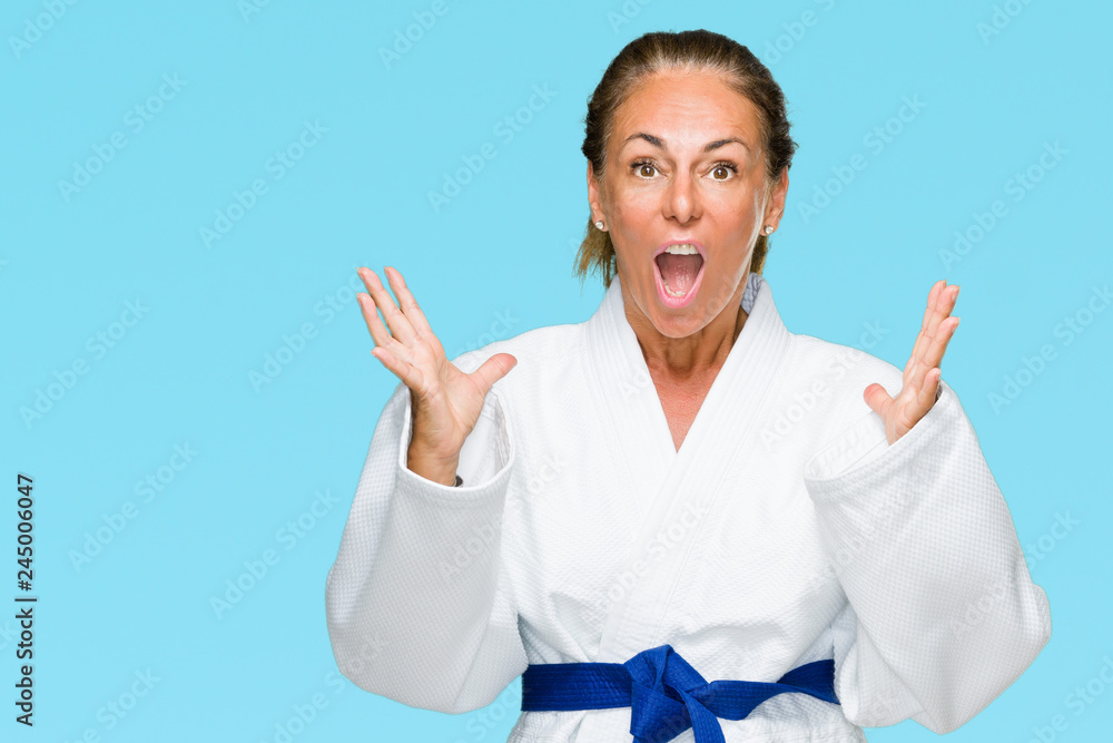 Middle age adult woman wearing karate kimono uniform over isolated background celebrating crazy and amazed for success with arms raised and open eyes screaming excited. Winner concept Middle age adult woman wearing karate kimono uniform over isolated background celebrating crazy and amazed for success with arms raised and open eyes screaming excited. Winner concept
