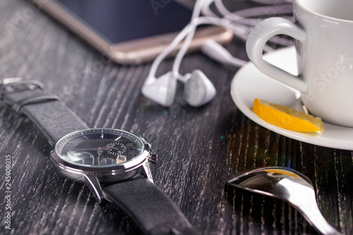 Wrist watch and mobile phone with headphones and a cup of coffee on a dark wooden table