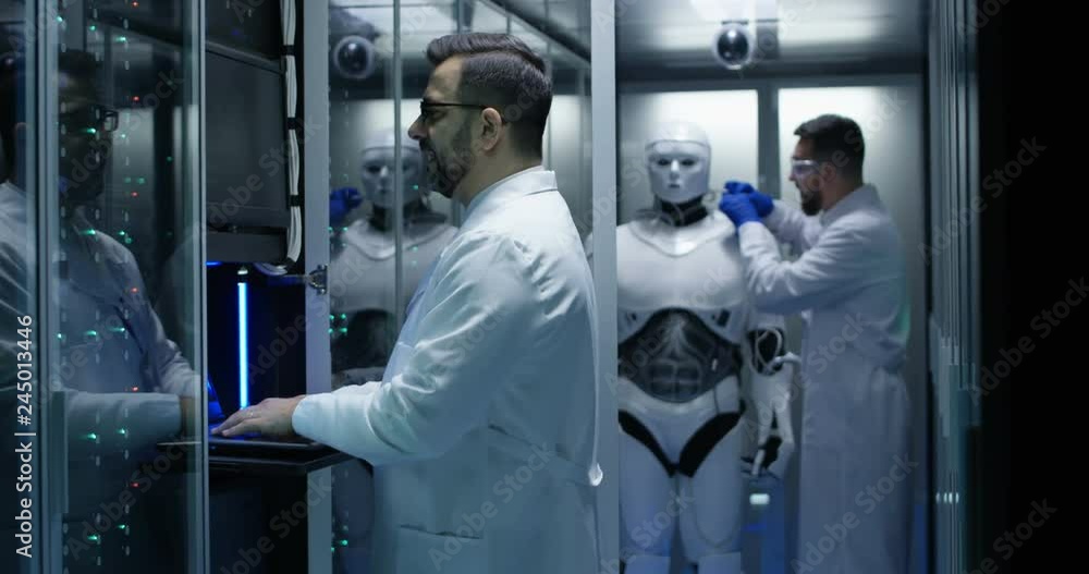 Medium shot of an engineer fixing wires of a robot while his colleague is testing controls on the tablet Medium shot of an engineer fixing wires of a robot while his colleague is testing controls on the tablet