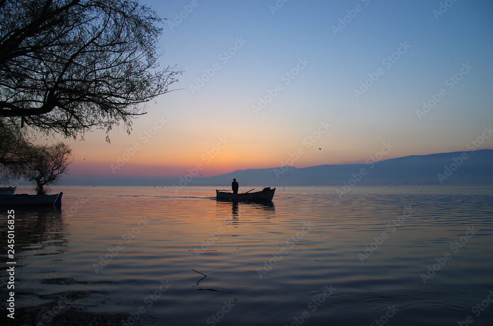 Golyazi, Bursa / Turkey - March 15 / 2014 : Silhouette of a fisherman staning on his boat while sunrise in the morning Golyazi, Bursa / Turkey - March 15 / 2014 : Silhouette of a fisherman staning on his boat while sunrise in the morning