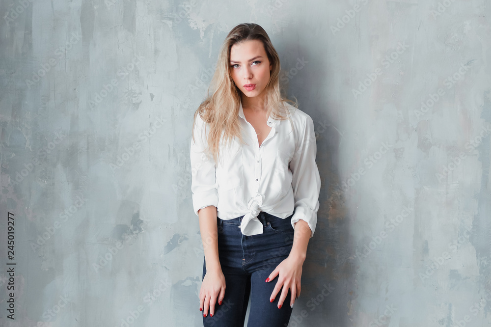 Model blonde blue jeans white shirt standing gray wall by window  copy space.