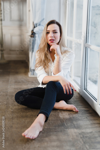 Model blonde white shirt blue pants jeans sitting on wooden parquet floor by window.
