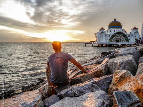 A man sitting in front of Malacca Strait Mosque, Malaysia. It is a World Heritage Site. Tender capture during the sunset, sun sets down in the sea. Solo traveler. 