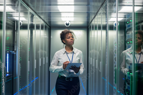 African American woman using tablet while walking in corridor of data center and checking hardware on server racks photo