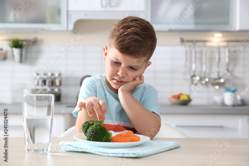 Unhappy little boy eating vegetables at table in kitchen photo