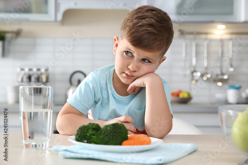 Unhappy little boy with plate of vegetables at table at table