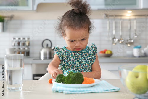 Cute African-American girl with plate of vegetables at table in kitchen