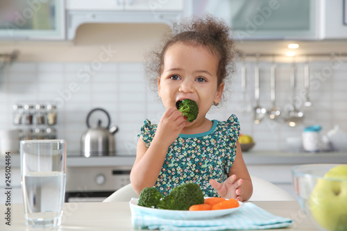 Cute African-American girl eating vegetables at table in kitchen
