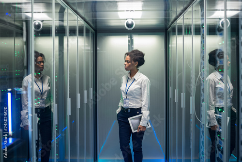 African American woman with tablet while walking in corridor of data center and checking hardware on server racks