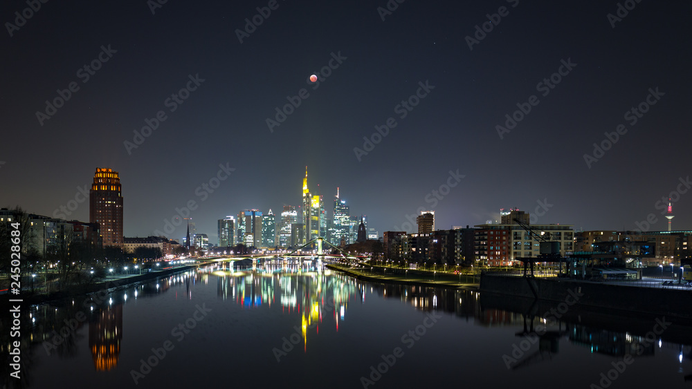 Blood moon at totality during lunar eclipse of over the skyline of Frankfurt, Germany, with reflections on the quiet river Main.
