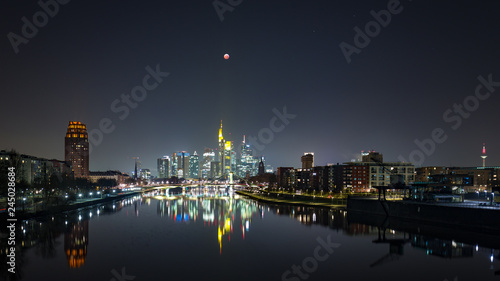 Blood moon at totality during lunar eclipse of over the skyline of Frankfurt  Germany  with reflections on the quiet river Main.