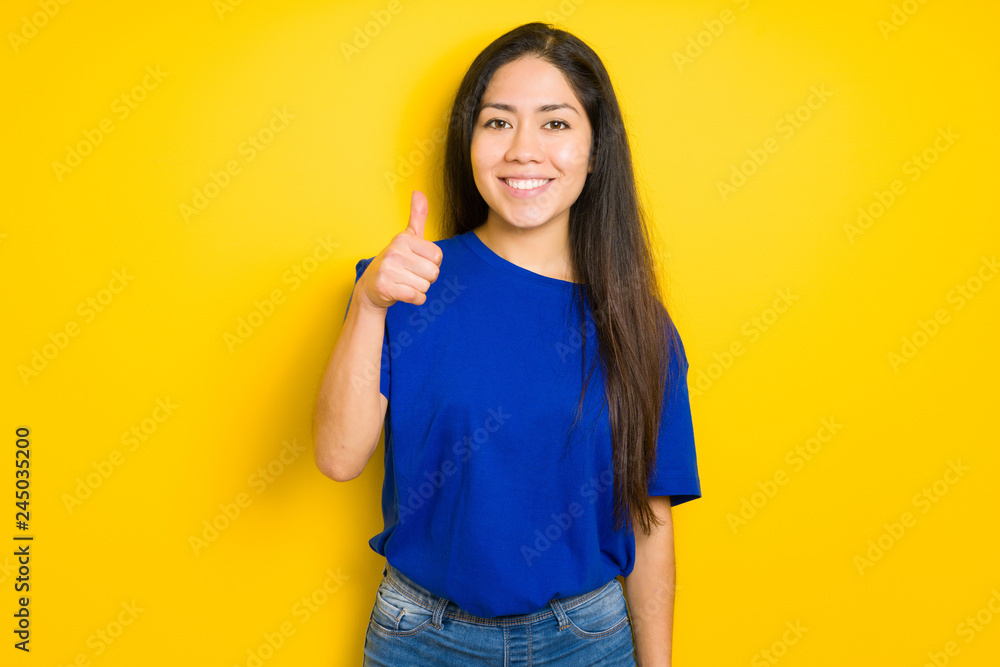 Beautiful brunette woman wearing blue t-shirt over yellow isolated background doing happy thumbs up gesture with hand. Approving expression looking at the camera with showing success. Beautiful brunette woman wearing blue t-shirt over yellow isolated background doing happy thumbs up gesture with hand. Approving expression looking at the camera with showing success.