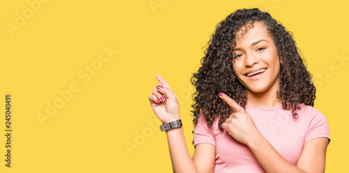 Young beautiful woman with curly hair wearing pink t-shirt smiling and looking at the camera pointing with two hands and fingers to the side.