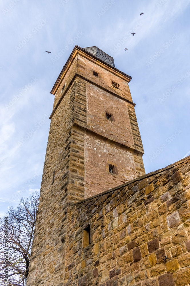 Wehrturm mit Zwiebeldach aus Schieferschindeln und Stadtmauer einer Stadtbefestigung aus Sandstein bei bedecktem Himmel und vier fliegende Vögel