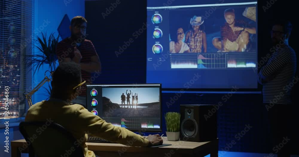 Medium shot of a young man editing video inside the modern video studio while his colleagues are giving instructions Medium shot of a young man editing video inside the modern video studio while his colleagues are giving instructions