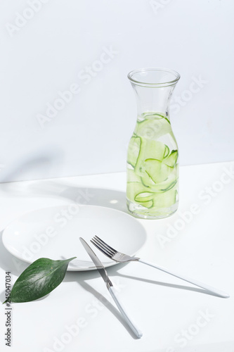 Plate, cutlery and jar of cucumber water on a white table. Creative minimalistic concept.
