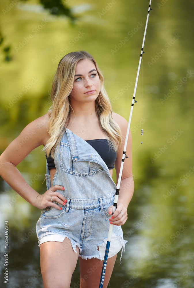 Beautiful blonde young woman fishing near creek wearing coveralls - looking  off Stock Photo