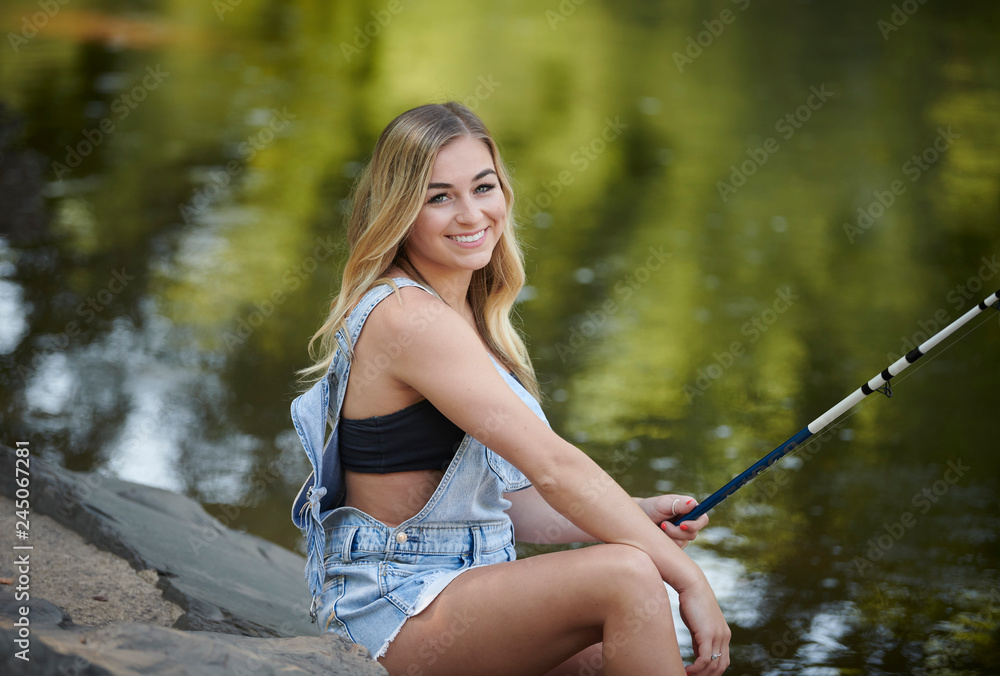 Beautiful blonde young woman fishing near creek wearing coveralls - seated  on rock Stock Photo