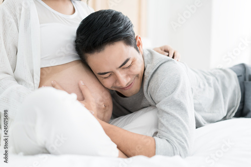 Asian Handsome man is listening to his Asian beautiful pregnant wife's tummy and smiling. Family love concept.