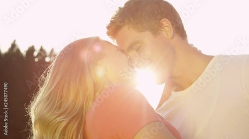 SLOW MOTION, SUN FLARE, CLOSE UP: Happy young woman and her smiling boyfriend rubbing their noses together while on a date in the beautiful sunny nature. Cheerful couple eskimo kissing in summer sun photo